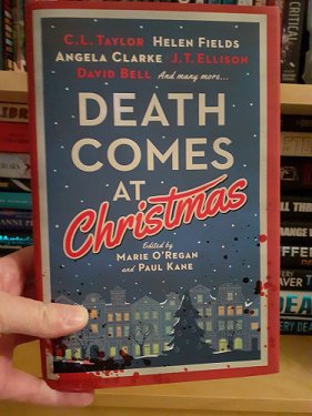 photograph of a hand holding a copy of Death Comes at Christmas, edited by Marie O'Regan and Paul Kane, up against full bookshelves