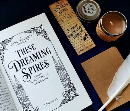 photograph of a copy of These Dreaming Spires, edited by Marie O'Regan and Paul Kane, open to the title page. Tol the right of the book are a These Dreaming Spires bookmark, an In These Hallowed Halls jar candle, a beige notebook and a white feather quill pen