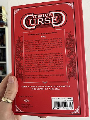 photograph of a man's hand holding a copy of the French edition of Twice Cursed, edited by Marie O'Regan and Paul Kane, up against a white background to show the back cover