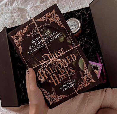 a hand holds a copy of In These Hallowed Halls, edited by Marie O'Regan and Paul Kane, tied with string, above a black box containing shredded black tissue paper, an Earl Grey teabag and an In These Hallowed Halls jar candle