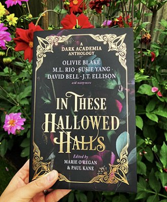 a hand holds a copy of In These Hallowed Halls, edited by Marie O'Regan and Paul Kane, in front of flowering bushes