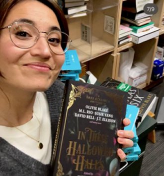 photograph of a smiling Olivie Blake holding up a copy of In These Hallowed Halls, edited by Marie O'Regan and Paul Kane