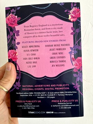 photograph of a man's hand holding a copy of The Hopeless Romantic's Guide to Enchantment, edited by Marie O'Regan and Paul Kane, up against a white surface to show the back cover. Contributing authors are listed: Kelley Armstrong, Olivia Atwater, A.Y. Chao, Vida Cruz-Borja, Alexis Hall, L.R. Lam, Hannah Nicole Maehrer, Juliet Marillier, Chloe Neill, Greer Stothers, Rebecca Thorne and Jen Williams