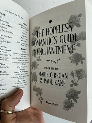 photograph of a man's hand holding up a copy of The Hopeless Romantic's Guide to Enchantment, edited by Marie O'Regan and Paul Kane, up against a white surface. The book is held open to show the interior title page
