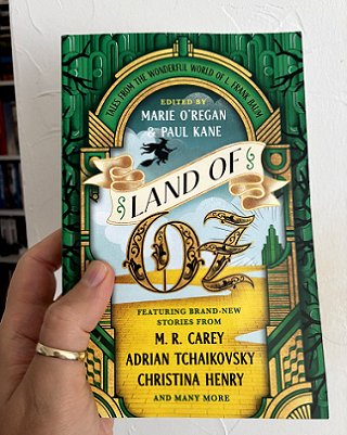 photograph of a man's hand holding up a copy of Land of Oz, edited by Marie O'Regan and Paul Kane, against a white background