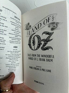 photograph of a man's hand holding a copy of Land of Oz, edited by Marie O'Regan and Paul Kane, up to show the interior title page
