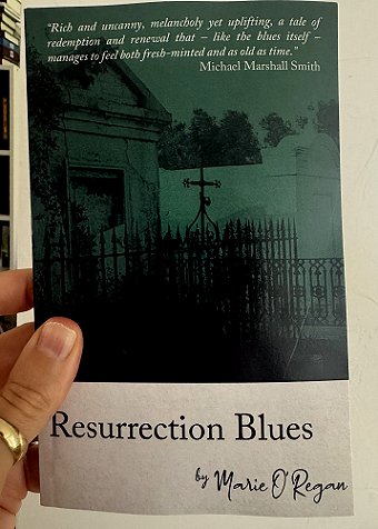 photograph of Paul's hand holding up a copy of Resurrection Blues by Marie O'Regan against a white background. Text reads Resurrection Blues by Marie O'Regan. At the top of the page of the following quote: Rich and uncanny, melancholy yet uplifting, a tale of redemption and renewal that - like the blues itself - manages to feel both fresh-minted and as old as time. Michael Marshall Smith