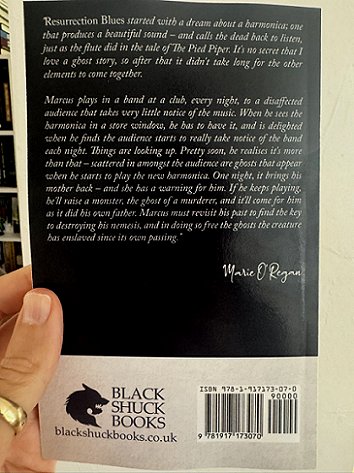 photograph of Paul's hand holding up a copy of Resurrection Blues by Marie O'Regan to show the back cover. Text reads: Resurrection Blues started with a dream about a harmonica, one that produces a beautiful dound - and calls the dead back to listen, just as the flute did in the tale of The Pied Piper. It's no secret that I love a ghost story, so after that it didn't take long for the other elements to come together. Marcus plays in a band at a club, every night, to a disaffected audience that takes very little notice of the music. When he sees the harmonica in a store window, he has to have it, and is delighted when he finds the audience starts to really take notice of the band each night. Things are looking up. Pretty soon, he realises it's more than that - scattered in amongst the audience are ghosts that appear when he starts to play the new harmonica. One night, it brings his mother back - and she has a warning for him. If he keeps playing, he'll raise a monster the ghost of a murderer, and it'll come for him as it did his own father. Marcus must revisit his past to find the key to destroying his nemesis, and in doing so face the ghosts the creature has has enslaved since its own passing. Marie O'Regan. Black Shuck Books. blackshuckbooks.co.uk