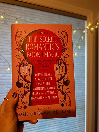 photograph of a woman's hand holding a copy of The Secret Romantic's Book of Magic, edited by Marie O'Regan and Paul Kane, against a window covered by a white blind and lit up fairy lights
