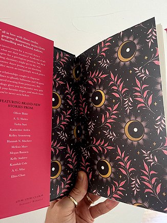 photograph of a man's hand holding up a copy of The Secret Romantic's Book of Magic, edited by Marie O'Regan and Paul Kane, to show the black endpapers, decorated with pink leaves, golden crescents and black circles with a white centre