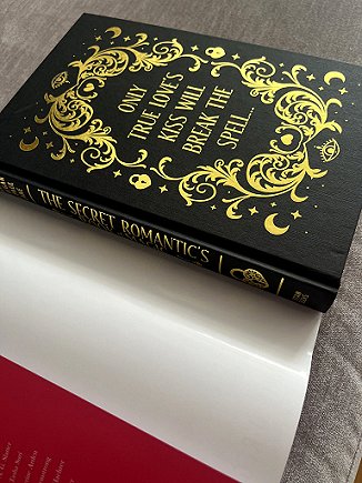 photograph of a copy of The Secret Romantic's Book of Magic, edited by Marie O'Regan and Paul Kane, lying on a grey surface. Dustjacket has been removed to show the black cover decorated with gold text and scrollwork, crescent moons, stars, locks and hearts. Text reads: ONLY TRUE LOVE'S KISS WILL BREAK THE SPELL...