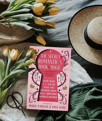 photograph of a copy of The Secret Romantic's Book of Magic, edited by Marie O'Regan and Paul Kane, lying on a beige fleece cloth, with a dark green scarf at the bottom righthand corner, and above that a straw hat with a black ribbon band and rim. On the left are two bunches of yellow tulips