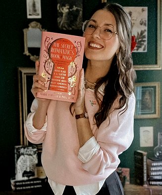 photograph of a smiling Kelly Andrew wearing a white shirt and pink sweater, holding up a copy of The Secret Romantic's Book of Magic, edited by Marie O'Regan and Paul Kane. Behind her is a desk with piles of books on top, and a dark green wall with several framed photographs and prints
