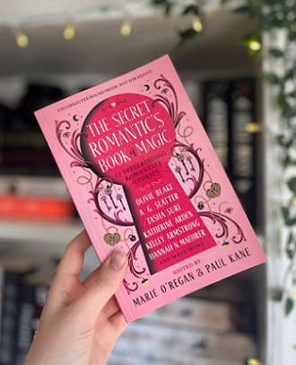 photograph of a hand holding a copy of The Secret Romantic's Book of Magic, edited by Marie O'Regan and Paul Kane, up against a white bookcase decorated with fairylights
