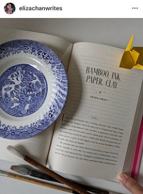 photograph showing a copy of The Secret Romantic's Book of Magic, edited by Marie O'Regan and Paul Kane, lying on a white surface; open at the title page of Eliza Chan's story: Bamboo, Ink, Paper, Clay. There are three paintbrushes beside the bottom of the book, and a piece of yellow origami at the top. A blue and white willow pattern dish is to the left.