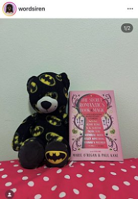 photograph of a copy of The Secret Romantic's Book of Magic, edited by Marie O'Regan and Paul Kane, standing against a white surface, on top of a pink cloth with white polka doats, beside a black teddy bear decorated with yellow and black batman symbols