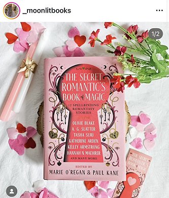 image of a copy of The Secret Romantic's Book of Magic, edited by Marie O'Regan and Paul Kane, on a white cloth scattered with pink heartshaped confetti. To the left of the book are pink and white candles tied with a gold ribbon. To the right is a bunch of deep pink flowers, and a Secret Romantic's bookamrk