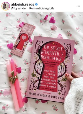 photograph of a woman's hand holding a copy of The Secret Romantic's Book of Magic, edited by Marie O'Regan and Paul Kane, aginst a white cloth decorated with pink flowers and scattered with pink heartshaped confetti. To the left of the book are two pink candles tied with a gold ribbon and a Secret Romantic's bookmark