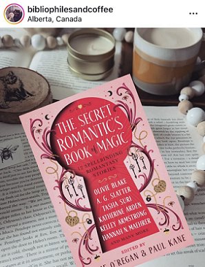 photograph of a copy of The Secret Romantic's Guide to Magic, edited by Marie O'Regan and Paul Kane, on top of an open book. To the right is a string of beige and white beads, to the left is a wooden coaster. At the top, from the left, more of the same beads, a candle in a brass tin, and a brown and white mug of coffee