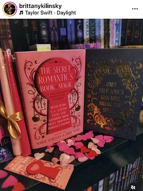 image showing a copy of The Secret Romantic's Book of Magic, edited by Marie O'Regan and Paul Kane, standing on a black surface in front of a row of books. The dustjacket stands beside the book, which has a black cover showing gold scrollwork surrounding text: ONLY TRUE LOVE'S KISS WILL BREAK THE SPELL.... To the left of the book are two pink candles tied with a gold ribbon, and a Secret Romantic's bookmark. In front of the book is pink heartshaped confetti