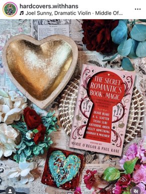 photograph showing a copy of The Secret Romantic's Book of Magic, edited by Marie O'Regan and Paul Kane, lying on a brown and cream platter atop pages from books. To the left is a gold coloured heartshaped bowl, and a bunch of red and white flowers. To the top and bottom right are more flowers in shades of red and pink, with dark and light green leaves, and at the bottom is a red square with a turquoise heart painted on it