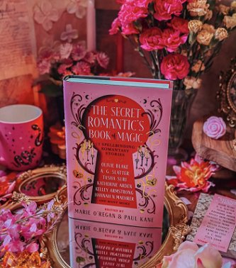 photograph of a copy of The Secret Romantic's Book of Magic, edited by Marie O'Regan and Paul Kane, standing on a circular gilt-edged mirror. To the left are pink flowers, a smaller handheld gilt-edged mirror and a pink mug with black snakes on it. To the right is a Secret Romantic's bookmark, and flowers in varying shades of pink