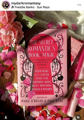 photograph showing a copy of The Secret Romantic's Book of Magic, edited by Marie O'Regan and Paul Kane, on top of a cream cloth, surrounded by pink heartshaped confetti. To the left are two pink roses, to the right are two pink candles tied with a gold ribbon
