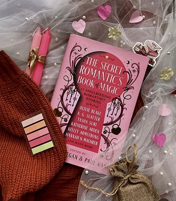 photo of a copy of The Secret Romantic's Book of Magic, edited by Marie O'Regan and Paul Kane, lying on a white veil studded with pearls, alongside two pink candles tied with a gold ribbon and a rust-coloured knitted hat to the left. On top of the hat are some post-it tabs ranging from white through orange, pink and yellow. On the right is a gold pouch and some pink heartshaped confetti