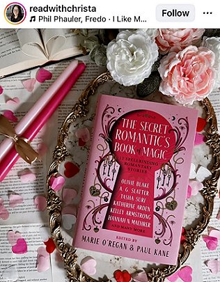image showing a copy of The Secret Romantic's Book of Magic, edited by Marie O'Regan and Paul Kane, lying on a gilt frame covered in pink and white heart-shaped confetti, on top of loose book pages. To the left are two pink candles tied with gold ribbon, and to the top are three roses - two pink, one white