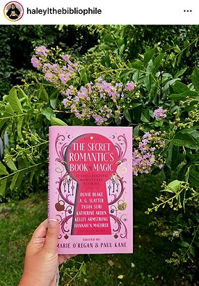 photograph of a hand holding a copy of The Secret Romantic's Book of Magic, edited by Marie O'Regan and Paul Kane, up against a bush with pale pink flowers