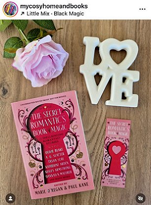 picture showing a copy of The Secret Romantic's Book of Magic, edited by Marie O'Regan and Paul Kane, lying on a wooden surface. To the right of the book is a matching Secret Romantic's bookmark. At the top left is a pink rose, and to the top right is a white ceramic ornament that spells LOVE