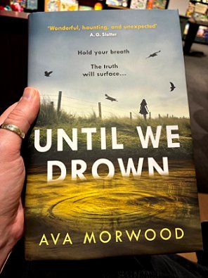 photograph showing a man's hand holding a copy of Until We Drown, by Ava Morwood