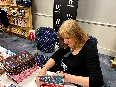 photograph of a smiling Marie O'Regan aigning a copy of The Secret Romantic's Book of Magic, edited by Marie O'Regan and Paul Kane, at the Waterstones table at WFC2025
